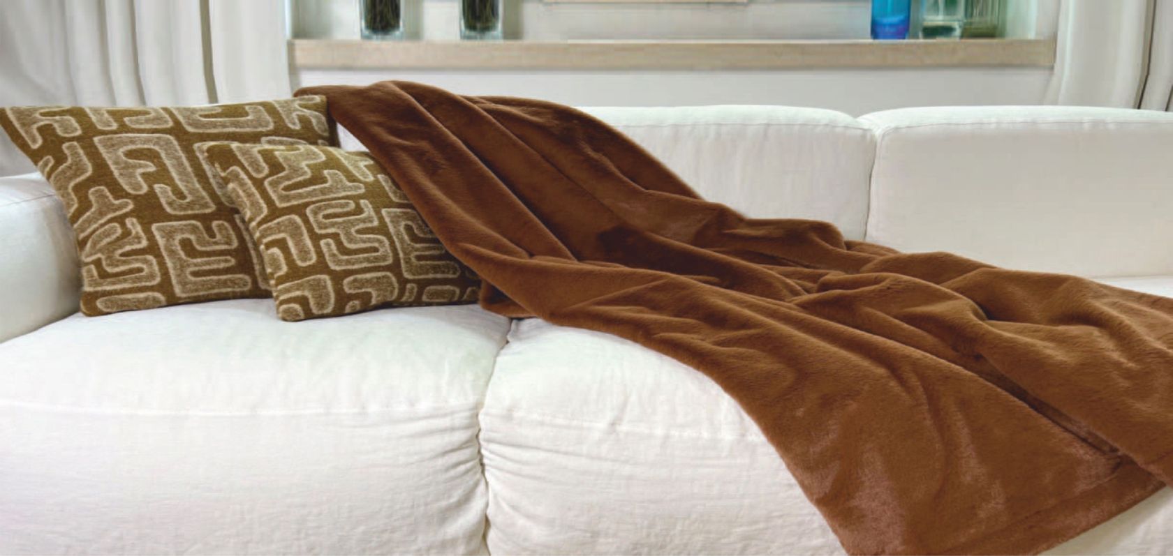 A Carma brown faux-fur throw draped across a white sofa with two patterned cushions in a styled living room, no people