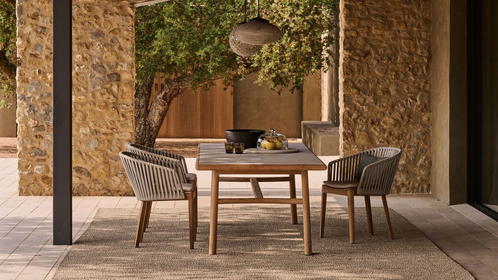 A Tribù teak dining table with woven-cord chairs under a stone loggia